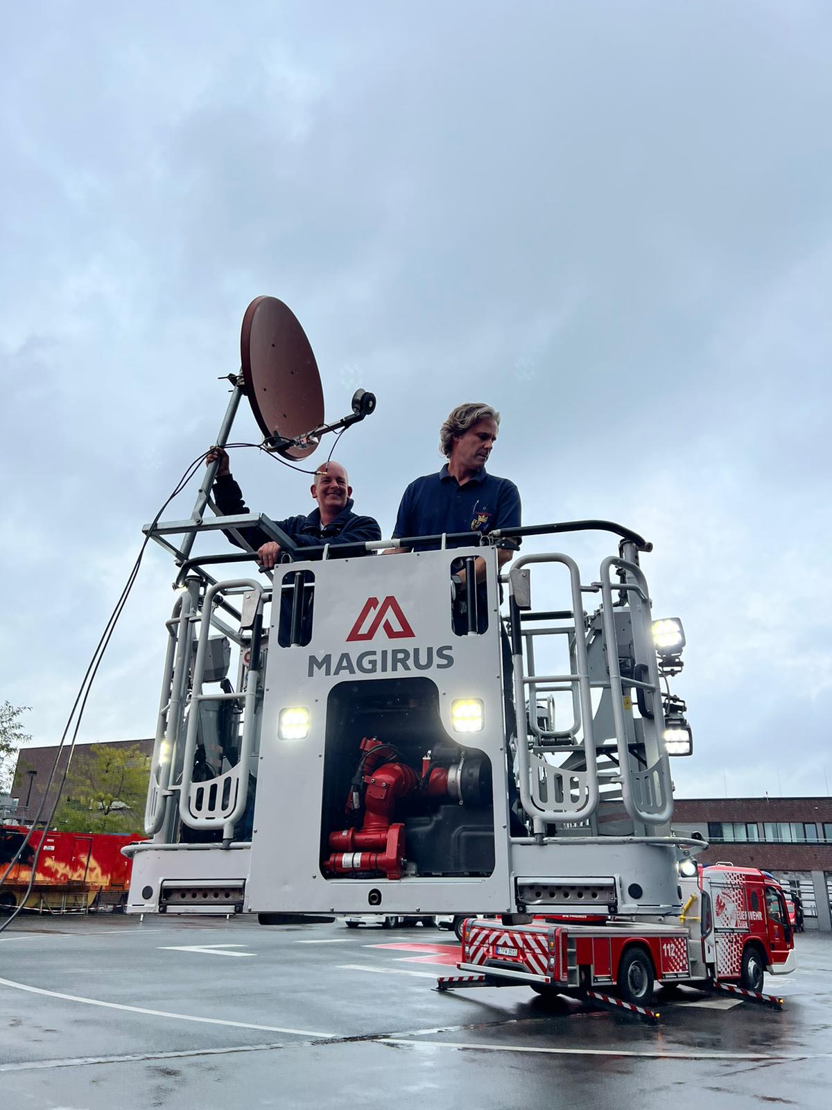 Zwei Personen befinden sich auf einer Feuerwehrdrehleiter. Eine Person steuert die Drehleiter, die andere hält eine Satellitenantenne in der Hand.